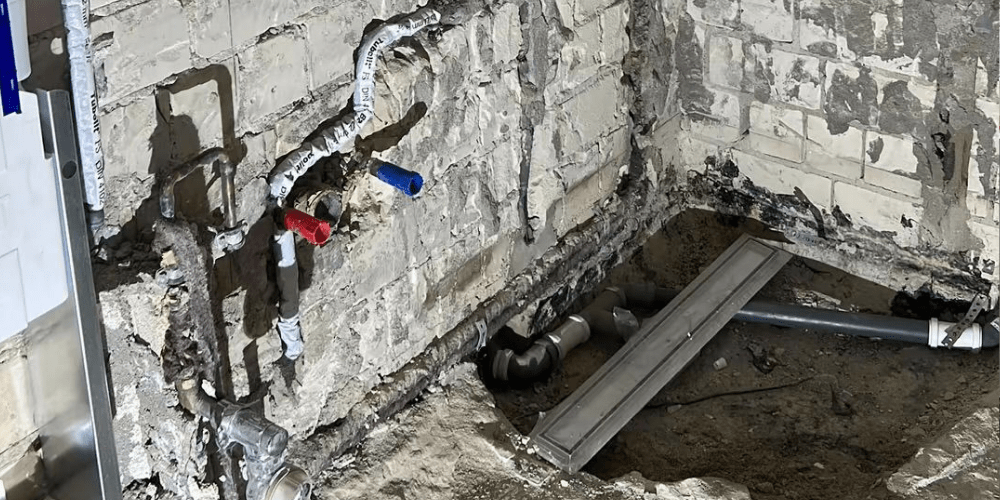 Exposed plumbing on a damaged basement wall with red, blue, and white pipes and a metal support beam nearby.