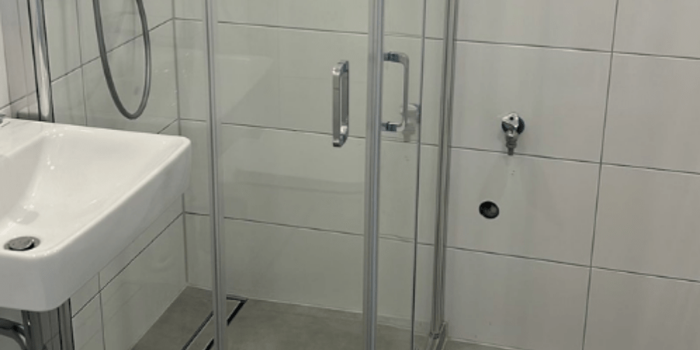 Bathroom interior featuring a glass-front shower enclosure, metal door handles, and a white wall-mounted sink beside tiled walls and a light gray floor.