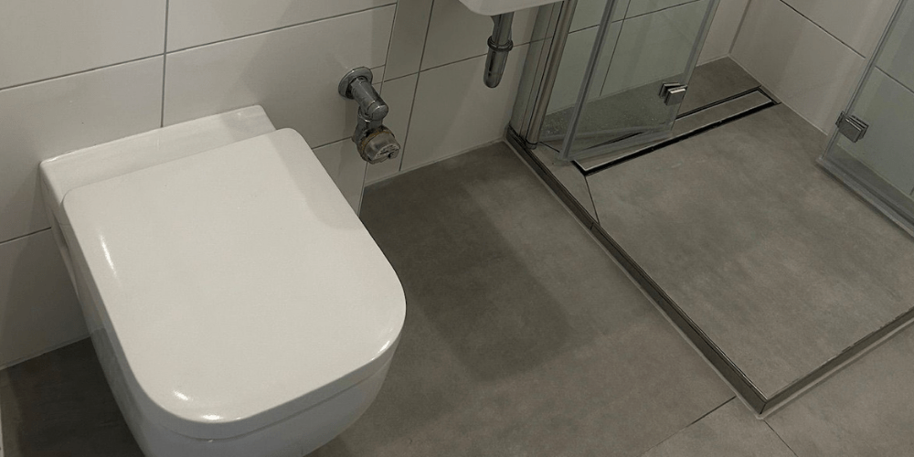White wall-mounted toilet beside a chrome pipe, with a glass-framed shower enclosure on a gray tiled bathroom floor.